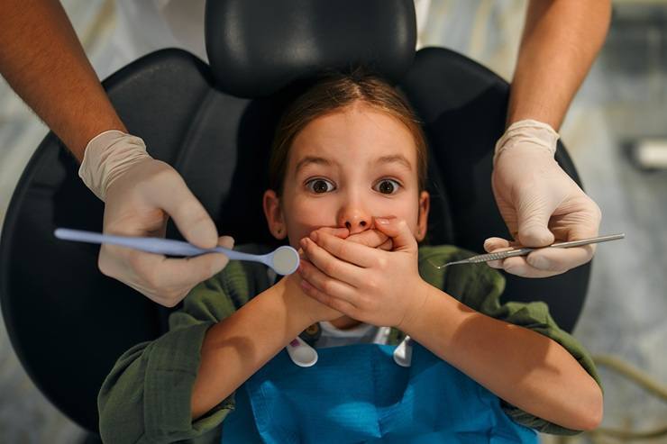 Child covering mouth in dental chair while dentist holds tools during examination.